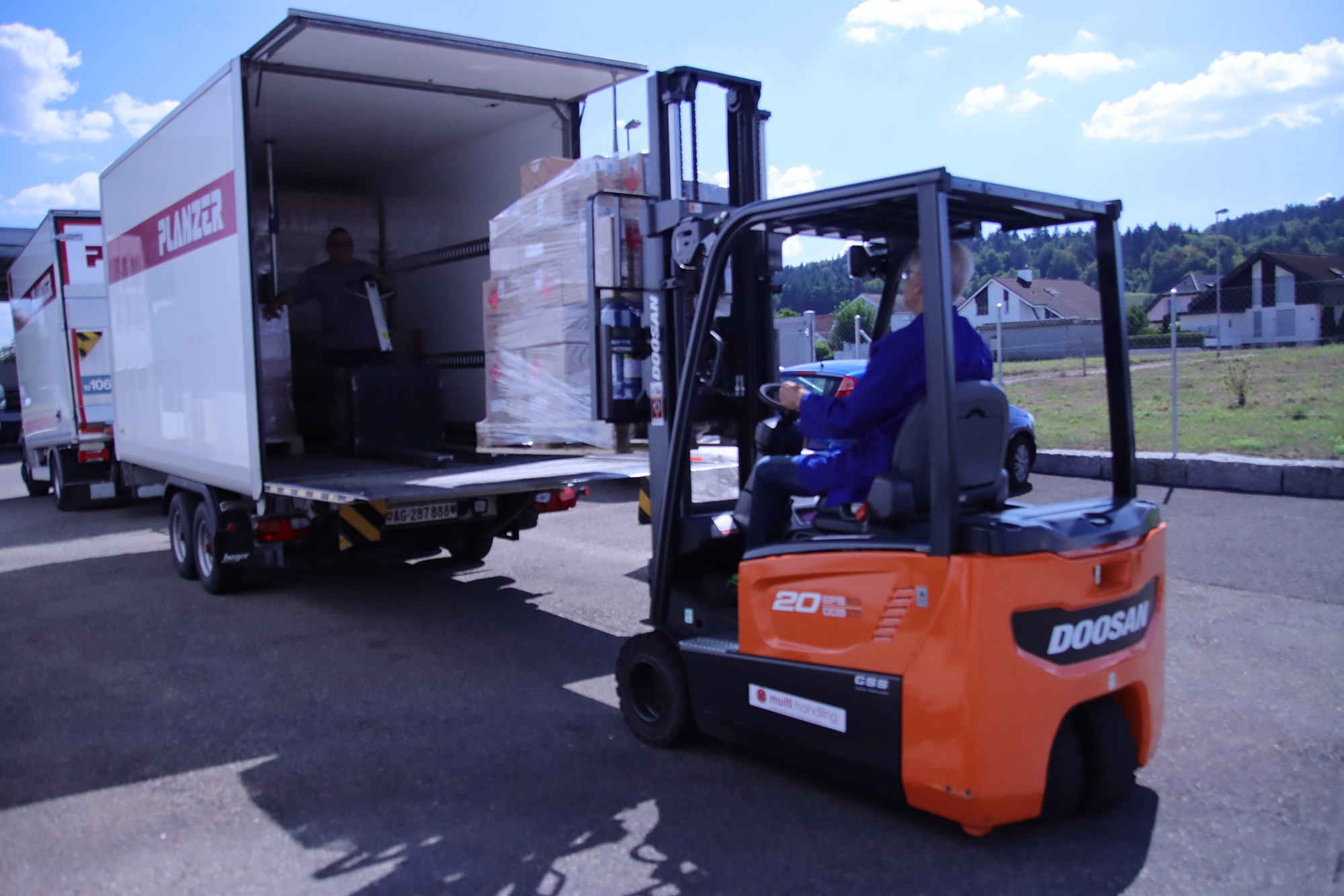Auf dem Bild ist ein grosser Doosan-Stapler erkennbar in orange, welcher abgepackte Chemischen Produkten in einen Planzer-Lastwagen verfrachtet.